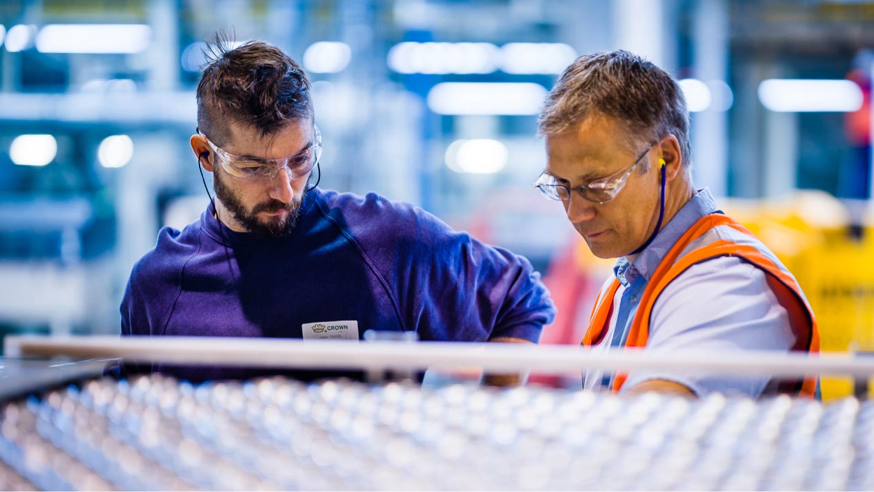 Two male crown employees in manufacturing type setting wtih safety googles, one younger man in blue sweatshirt, one older gentleman in blue collared shirt sleeve button down with safety vest