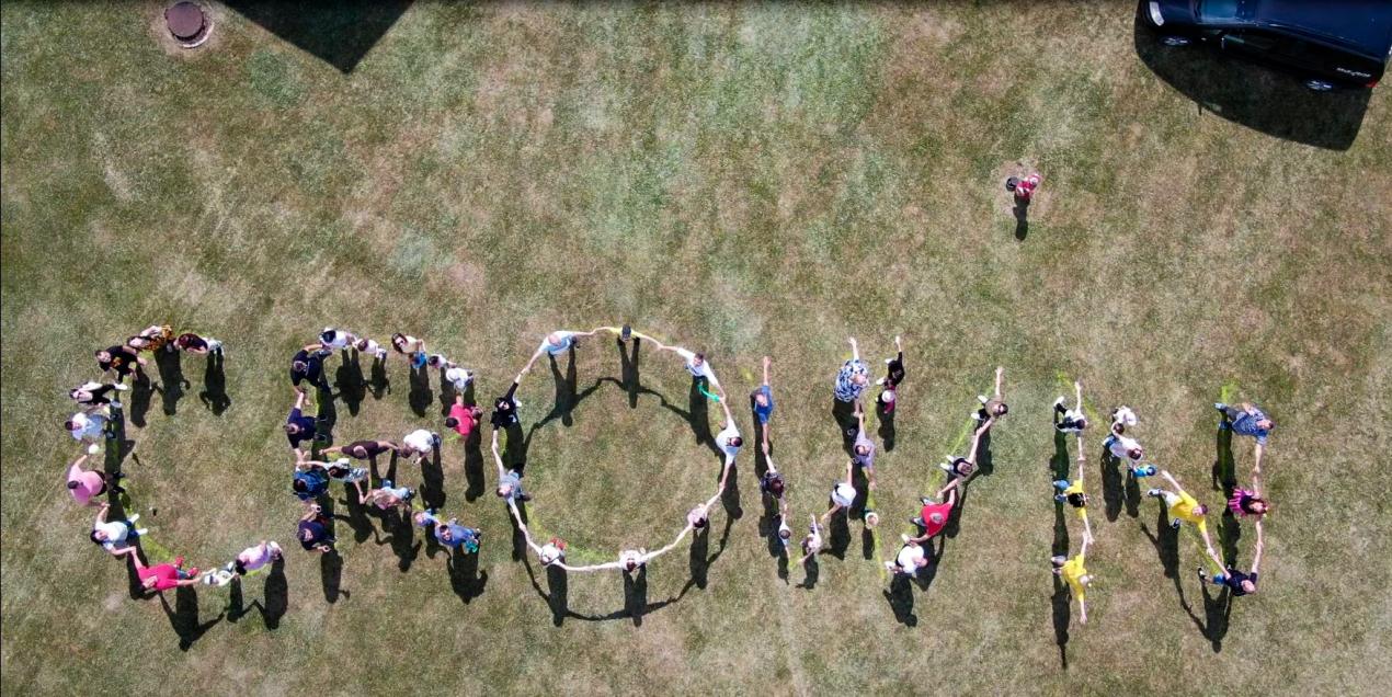 A large group of people spell out the word CROWN against a backdrop of green and brown grass.