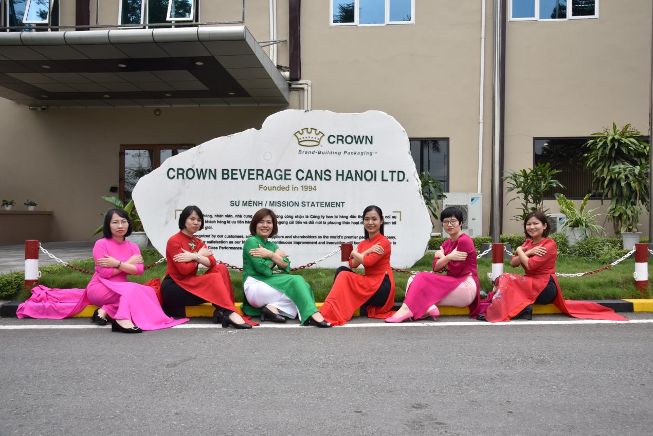A group of Asian women sit outside in front of a Crown Cork sign. They have their arms crossed in the symbol for "equity".
