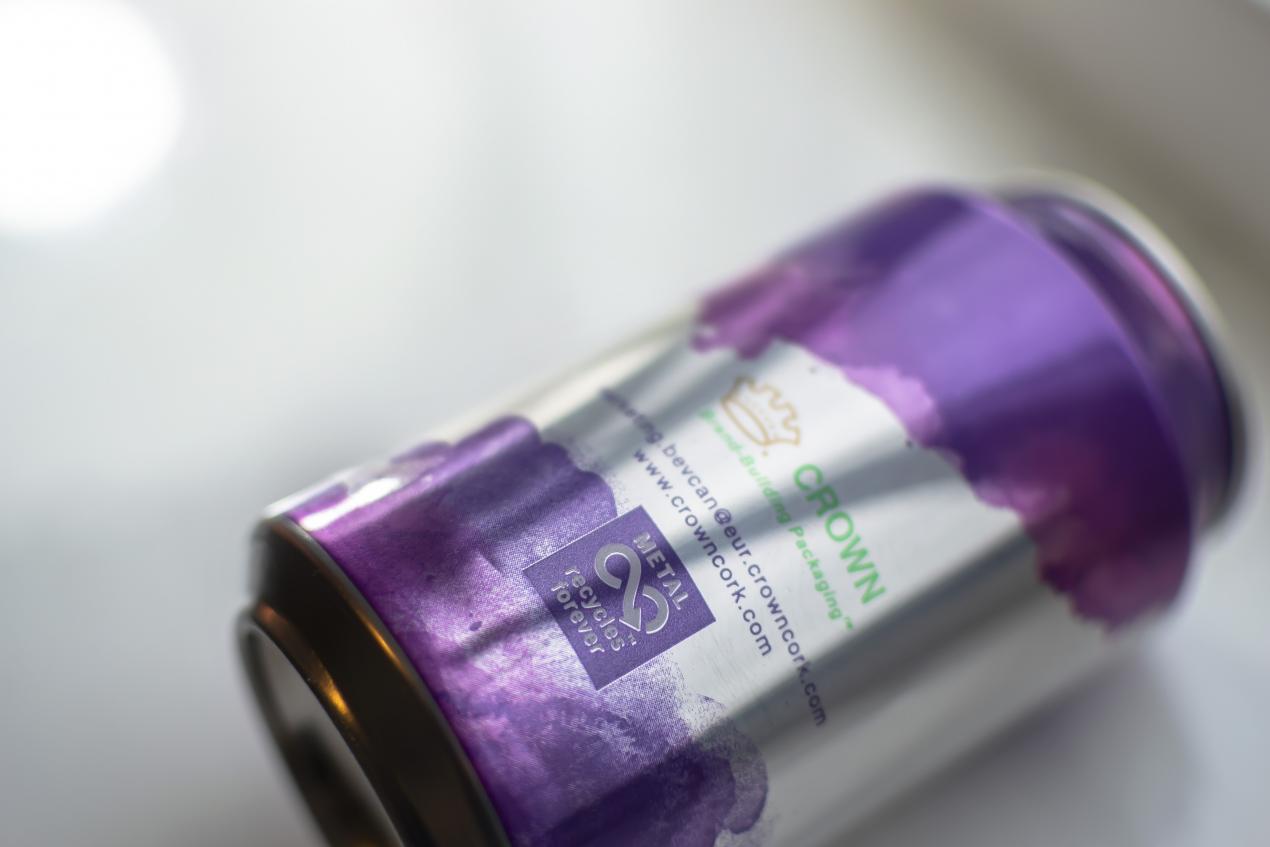 A Crown aluminum can with purple watercolor on the top and bottom lays against a white background. It says "metal recycles forever" on the side.