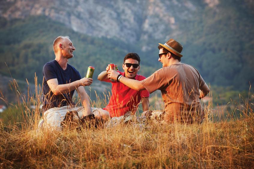 Several male-presenting people sit outdoors and clink glasses together.