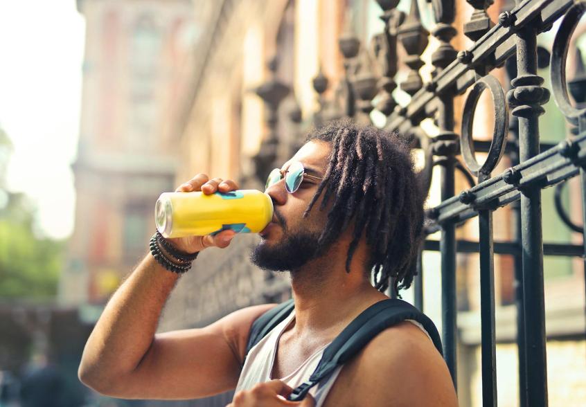 A male-presenting person drinks out of an aluminum can outdoors.